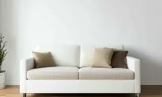A photograph depicts a minimalist living room interior. The composition centers on a white sofa with a beige cushion and brown throw pillows. The sofa is positioned against a plain white wall. Natural light streams in from an unseen window, casting a soft shadow on the wall. The floor is whitewashed wood. The overall aesthetic is calm, serene, and modern, with a muted color palette of white, beige, and brown. The image evokes a feeling of tranquility and simplicity. There are no synthetic elements visible. The lighting is soft and diffused, creating a gentle atmosphere. The style is Scandinavian minimalist. The vibe is peaceful and relaxing.